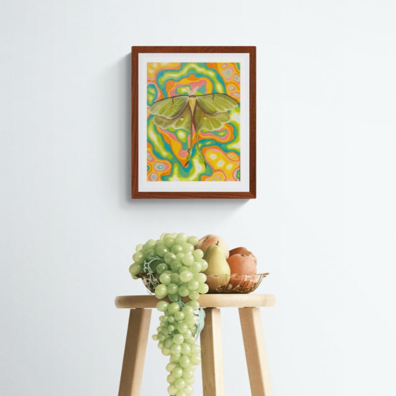 Framed artwork of a colorful lunar moth on a white wall above a wooden stool with grapes and apples.