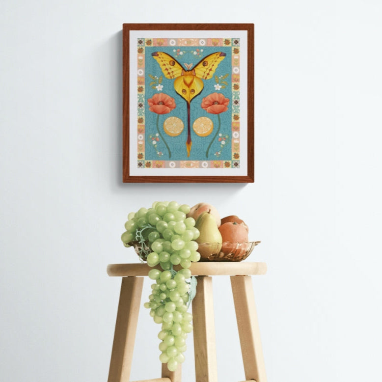 Framed artwork of a comet moth and flowers on a white wall above a wooden stool with fruit and grapes.